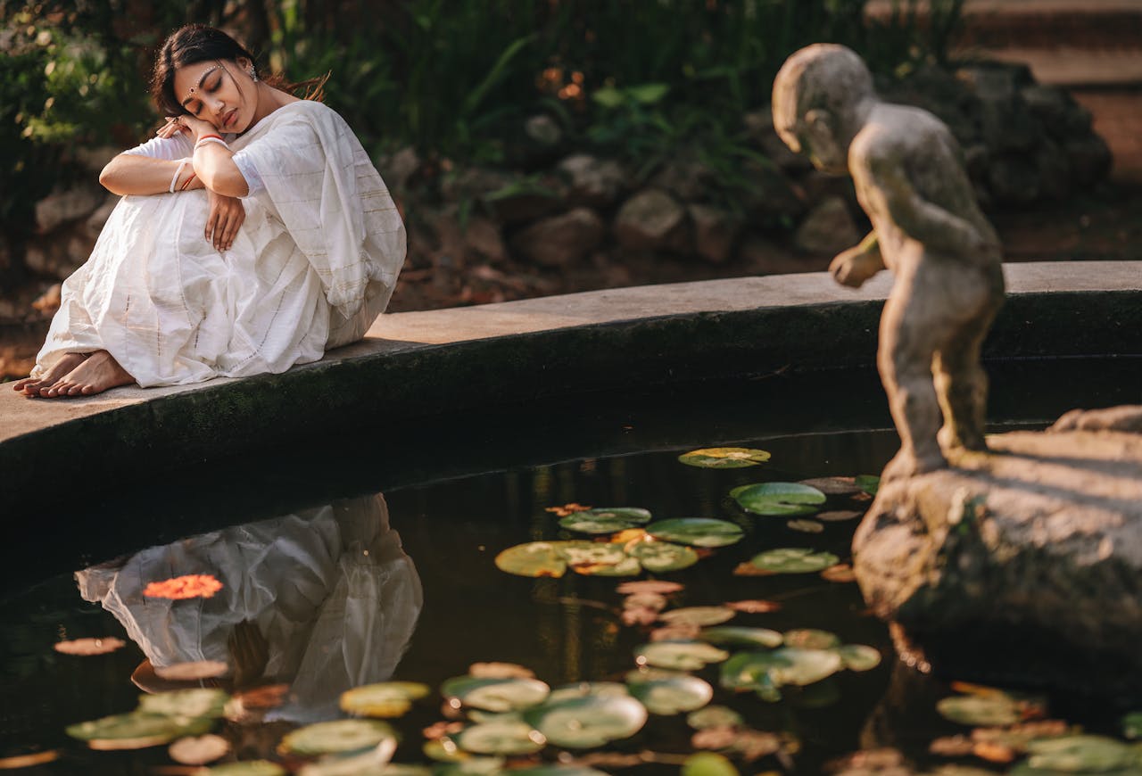 Woman in a white saree resting by a peaceful pond with a statue, reflecting tranquility and beauty.