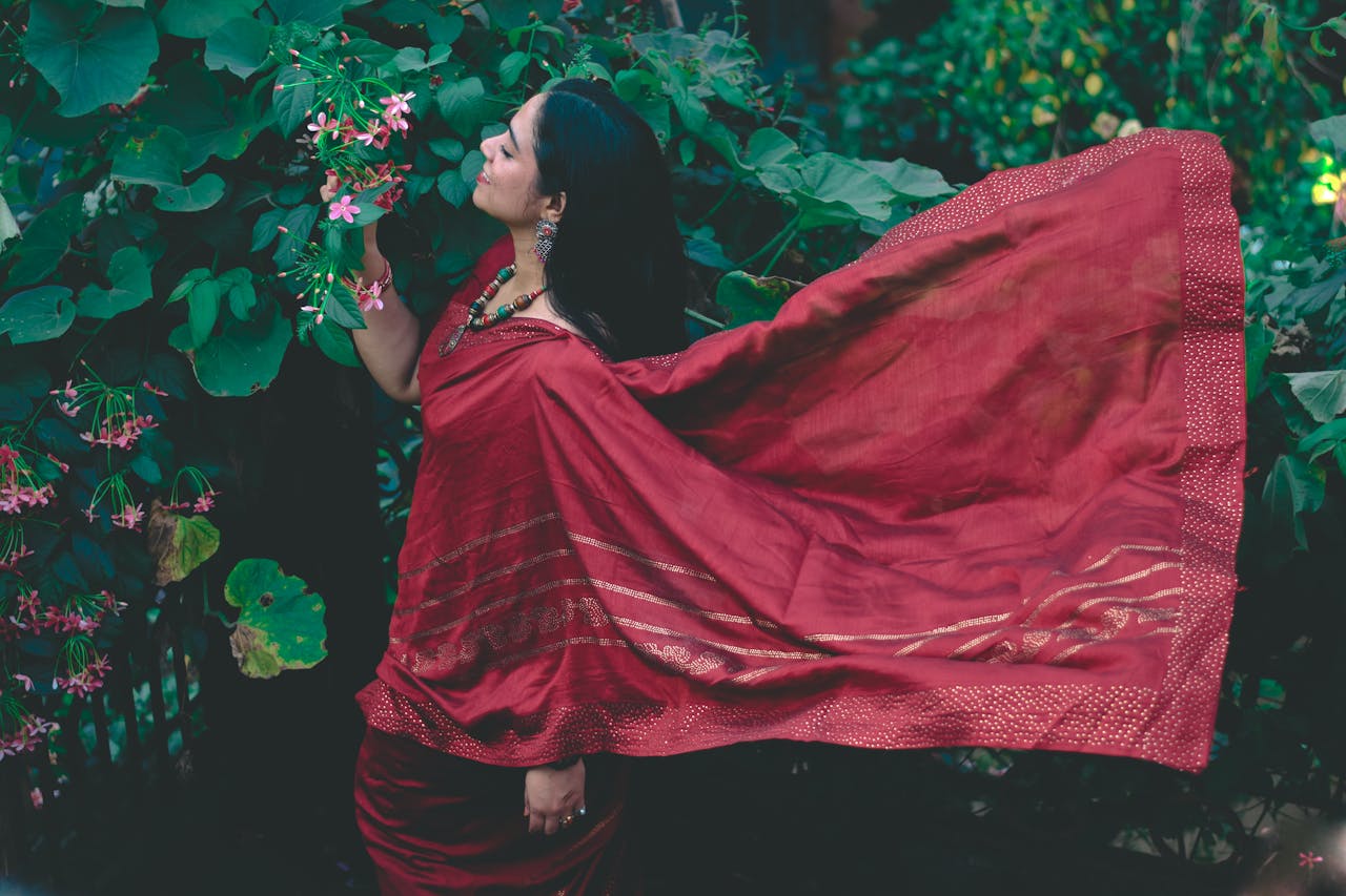 services-01 Woman in a flowing red saree stands among lush green foliage, embracing traditional Indian beauty.