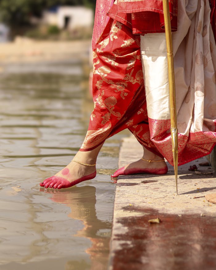services-02 A woman stands near a riverbank wearing a traditional red saree and anklets, touching water.