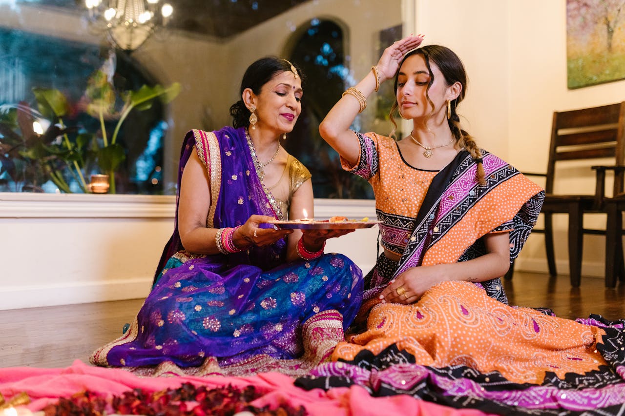 services-03 Mother and daughter performing a traditional Indian ritual indoors, wearing colorful sarees.
